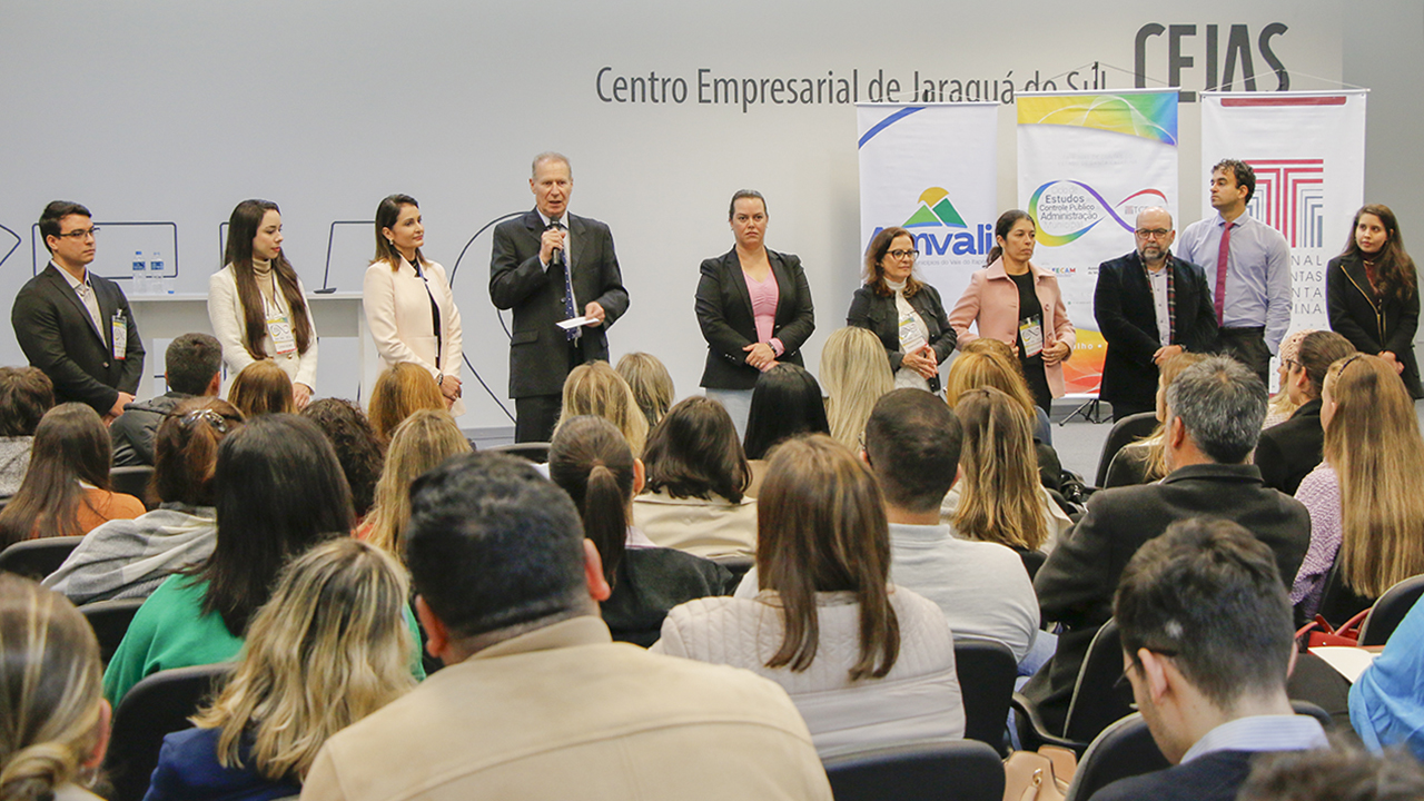 Foto do Presidente do TCE/SC, conselheiro Herneus De Nadal, falando ao microfone durante a etapa de Jaraguá do Sul do Ciclo. Ele é um homem branco, de cabelos claros, alto, vestindo terno preto. Ao lado dele, há outras nove pessoas que o acompanham. Há também o banner do Ciclo de Estudos, do TCE/SC e da Amvali ao fundo.