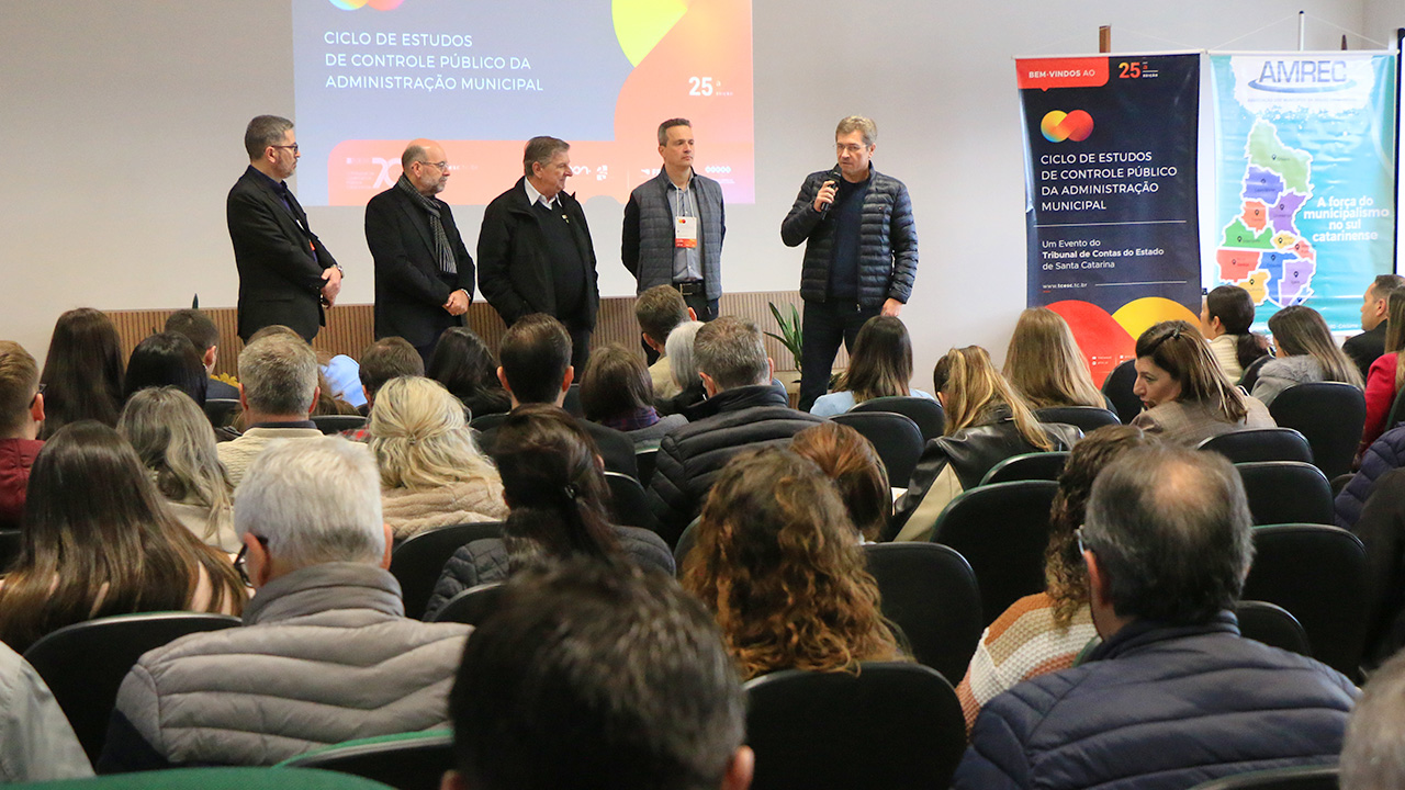 Banner horizontal com foto da equipe do TCE/SC no palco durante etapa de Lauro Müller do Ciclo de Estudos. Há cinco homens, um ao lado do outro. Entre eles, o vice-presidente do TCE/SC, José Nei Ascari, e o diretor do Icon, Moisés Hoegenn. À frente deles, de costas para a foto, a plateia participante. Ao fundo, no telão, projeção do logo do evento.