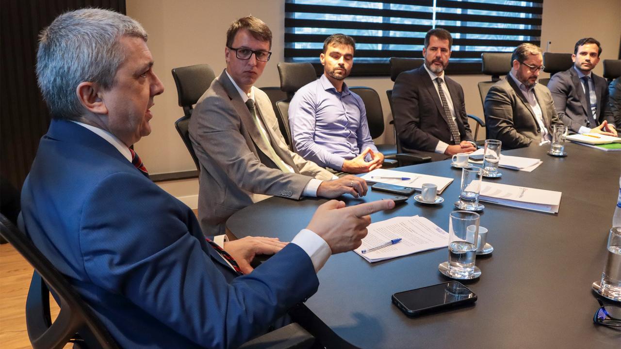 Banner horizontal com foto das autoridades durante reunião da mesa de consensualismo. Entre os presentes, o coordenador da mesa, auditor Márcio Medeiros, o defensor publico-geral, Ronaldo Francisco, e integrantes das demais instituições participantes. Eles estão sentados diante de uma mesa, enquanto conversam.