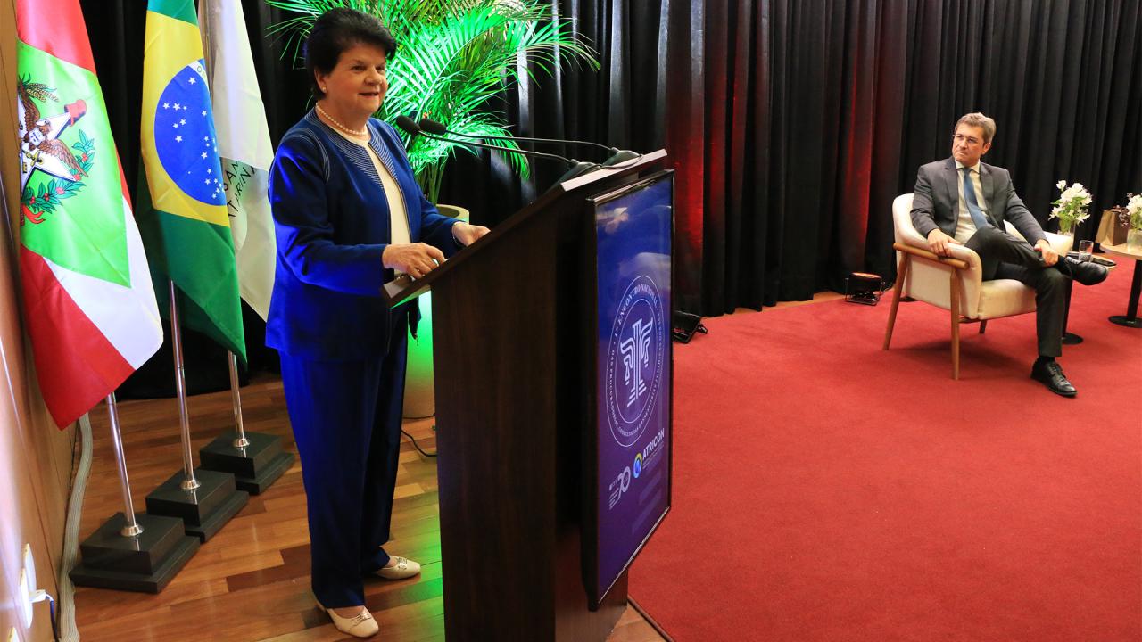 Foto horizontal ampla que mostra a palestrante Maria Sylvia Di Pietro falando ao público no púlpito do auditório do TCE/SC. Ela é uma mulher branca, de cabelos pretos e curtos. Veste roupa azul. Ao lado, em segundo plano, o conselheiro José Nei Ascari, mediador da palestra.