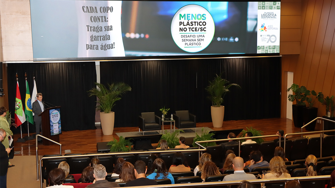 Banner horizontal com foto ampla do conselheiro José Nei Ascari discursando na abertura do evento, no palco do auditório do TCE/SC. Atrás dele, um telão que projeta o selo do desafio “Menos plástico no TCE/SC”. À frente dele, várias pessoas sentadas em poltronas, assistindo à sua fala. 