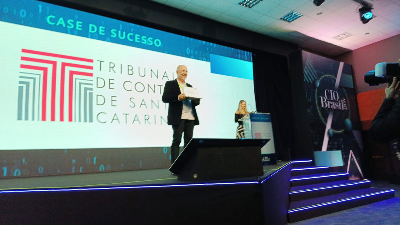 foto com o assessor da Aget, Jairo Wensing. Ele está em pé, ao centro, e segurando o certificado. Ao fundo, no telão, está projetada a logo do TCE/SC.