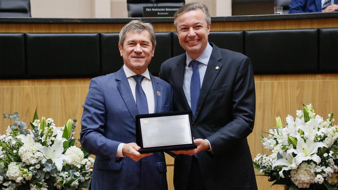 Foto horizontal do vice-presidente do TCE/SC, conselheiro José Nei Ascari, ao lado do deputado Napoleão Bernardes durante a sessão especial. Ambos sorriem, vestem terno e gravata e seguram, nas mãos, a homenagem, em formato de placa, recebida pelo conselheiro Ascari.