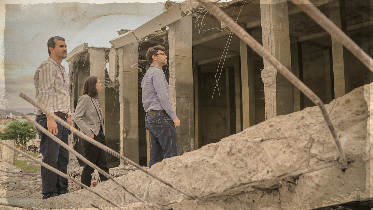 Foto do dia do acidente, em sépia para caracterizar que não é de agora, mostra três auditores do TCE/SC observando uma estrutura caída de concreto e ferros. Os ferros aparecem em primeiro plano.