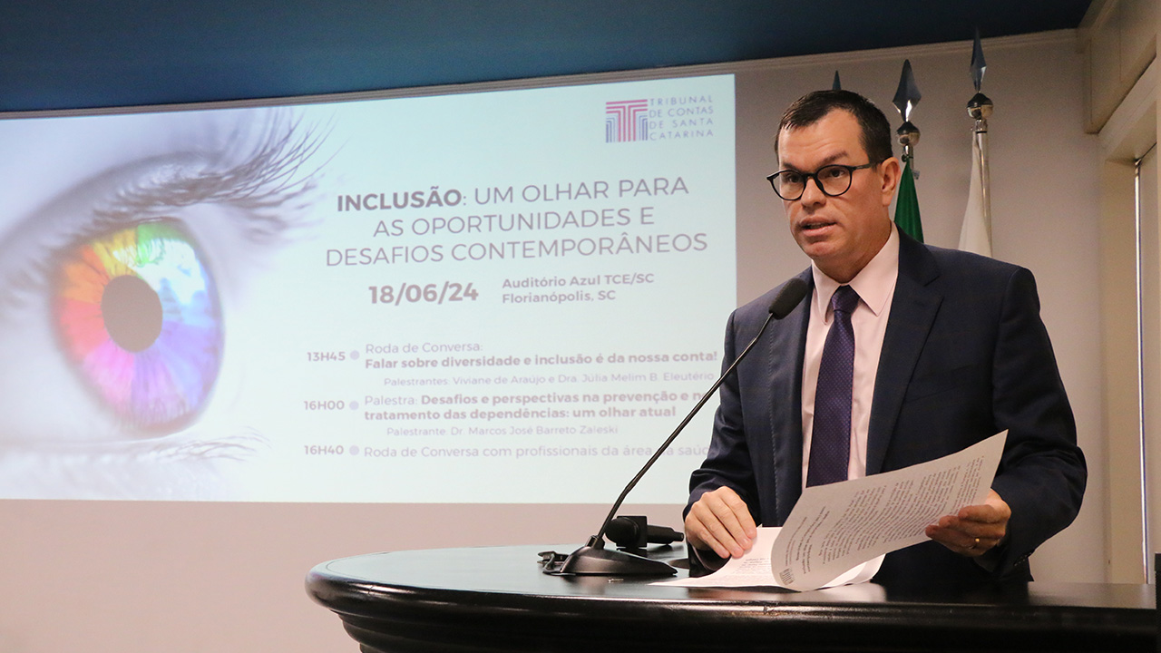 Banner horizontal com foto do conselheiro Adircélio de Moraes Ferreira Júnior durante a abertura do evento. Ele é um homem branco, de cabelos pretos. Veste terno e gravata e usa óculos de grau. Está em pé, diante do púlpito, falando ao microfone. Ao fundo, no telão, são projetados o logo e as informações do evento. 