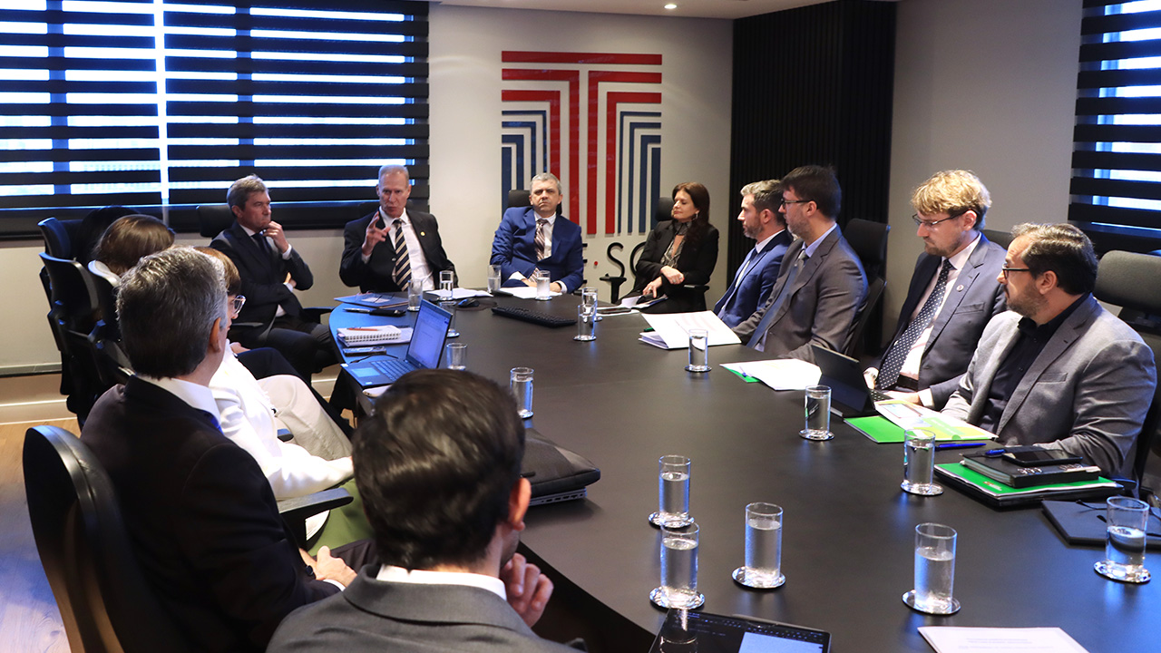 Banner horizontal com foto de várias pessoas sentadas ao redor de uma mesa durante reunião. Entre os presentes, o presidente do TCE/SC, conselheiro Herneus De Nadal, que está na ponta e fala aos demais, o conselheiro José Nei Ascari, ao seu lado, e representantes da DPE/SC, da OAB-SC, da SEF e de outras instituições. Ao fundo, na parede, plotagem do logo do Tribunal de Contas.