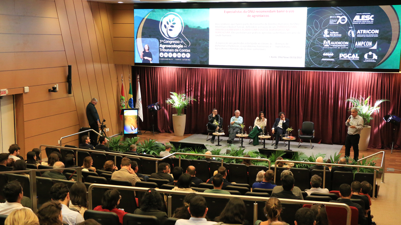 Banner horizontal com foto de um dos painéis, no auditório do TCE/SC, durante o congresso. Em destaque, participantes acompanham a programação sentados na plateia, lado a lado, enquanto, no palco, painelistas participam de debate e um palestrante fala ao público com microfone. Ao fundo, grande telão exibe a identidade visual do evento, conteúdo da apresentação e logos de instituições parceiras.