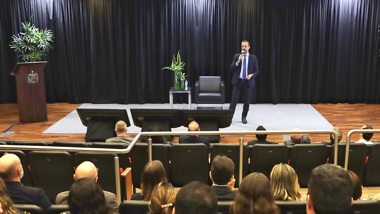 Imagem mostra palestrante no centro de um palco composto por tapete cinza e uma poltrona preta, com fundo preto. Em primeiro plano, pessoas de costas assistindo à apresentação 