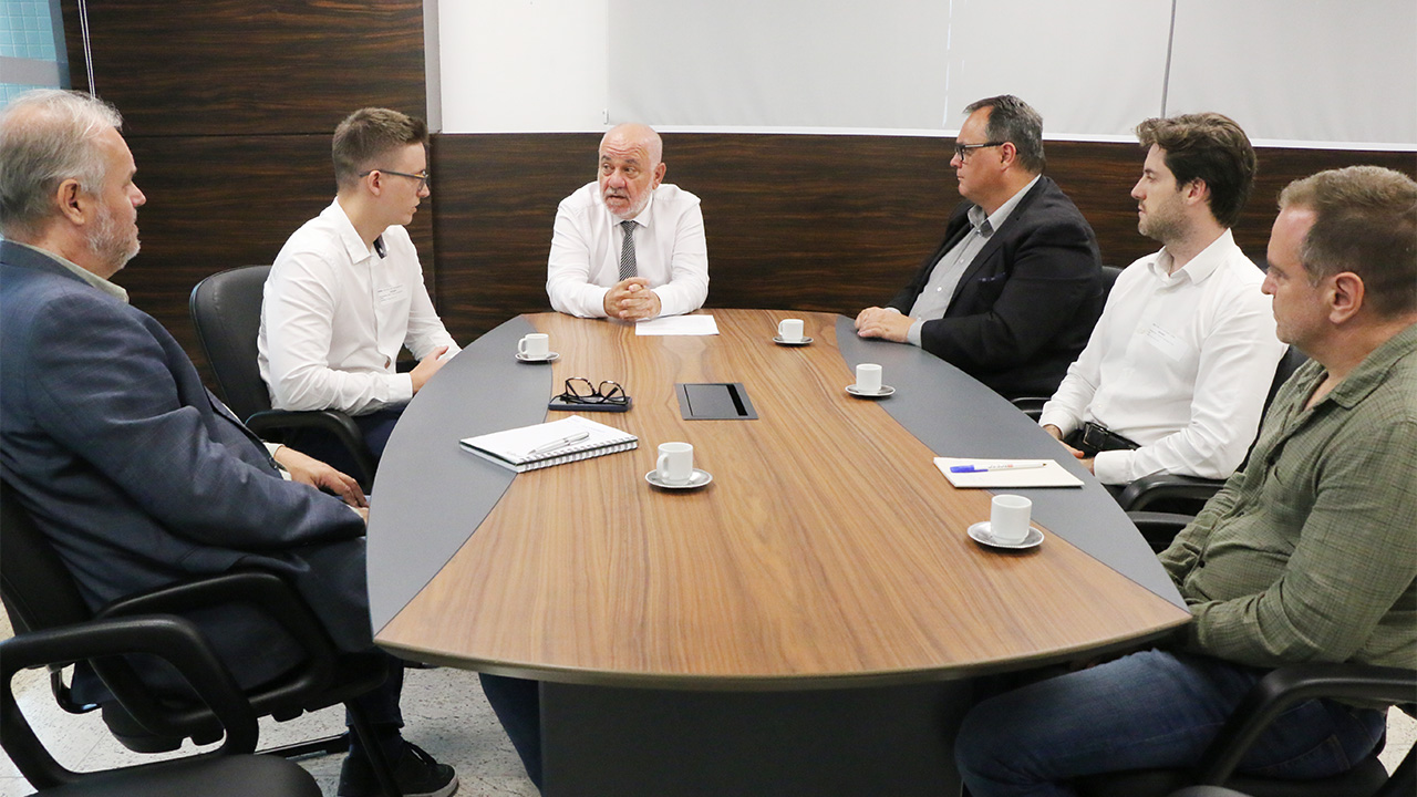 Imagem mostra seis pessoas reunidas em torno de uma mesa com tons de madeira e preto