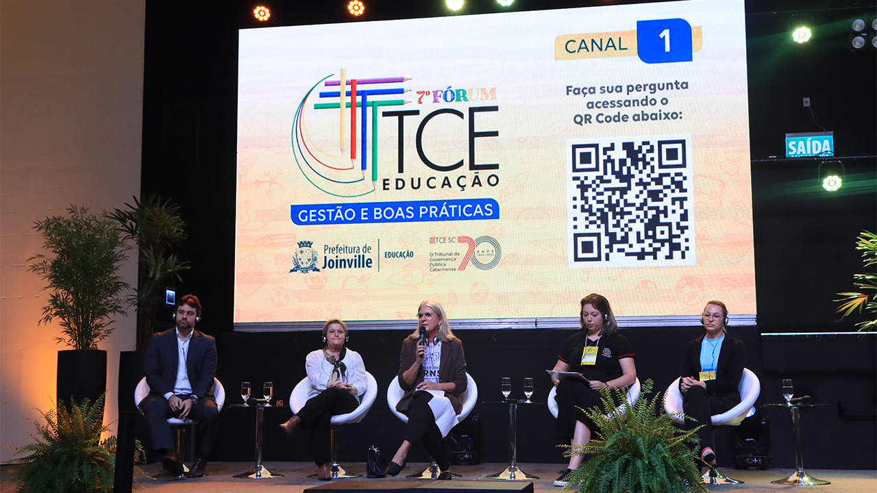 Imagem mostra cinco pessoas sentadas em poltronas brancas à frente de um telão que mostra o logotipo do Fórum TCE Educação, com predominância do tom amarelo. Há um homem, à esquerda, e quatro mulheres na imagem