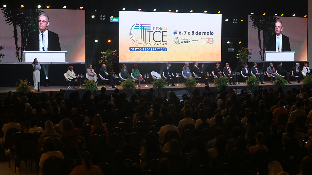 Foro mostra um palco com 14 pessoas sentadas uma ao lado da outra, de frente para a plateia. Ao fundo há três telões. O do centro mostra a logomarca do TCE Educação e do evento. Os laterais mostram um homem falando ao microfone.