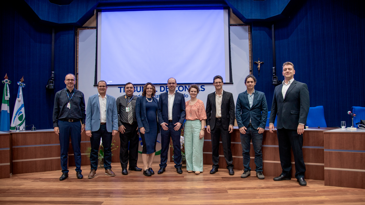 Foto do auditório do TCE/MS, onde aparecem nove pessoas, de pé, enfileiradas, entre elas, o diretor Nilsom Zanatto e os demais técnicos dos dois tribunais. Ao fundo, um telão sem imagens, em branco. E no canto esquerdo as bandeiras do Estado do Mato Grosso do Sul e do TCE/MS. 
