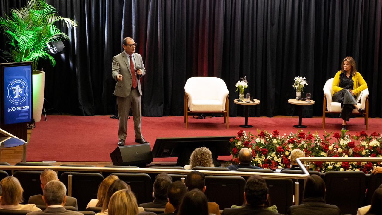 Foto horizontal que mostra o doutor em Direito Rodrigo Valgas palestrando no palco do auditório do TCE/SC. Ele está em pé, ao lado da mediadora, procuradora-geral Cibelly Farias. À sua frente, de costas, aparece parte do público participante, sentado em poltronas.