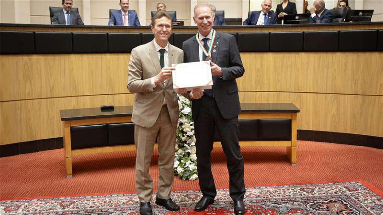 Foto horizontal posada do deputado Volnei Weber ao lado do conselheiro Herneus De Nadal, que tem uma medalha no pescoço e segura o certificado recebido, em sessão na Alesc. Ambos vestem terno e gravata e sorriem.