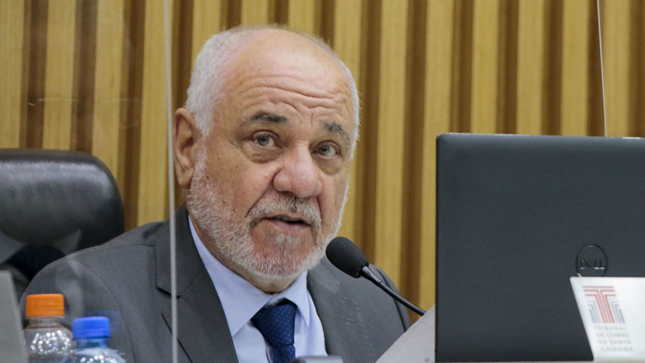 Foto do conselheiro Luiz Eduardo Cherem no Pleno do TCE/SC. Ele é um homem branco, de cabelos e barba grisalhos e de olhos claros. Está vestindo terno cinza, camisa branca e gravata azul, e falando ao microfone. 