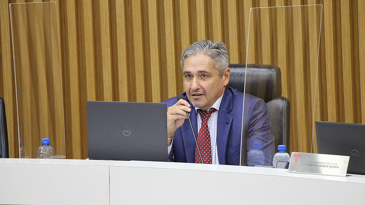  Foto do conselheiro Aderson Flores no Pleno do TCE/SC. Ele é um homem branco, de cabelos grisalhos. Veste um terno azul-escuro e gravata vermelha. Está sentado diante de um computador e falando ao microfone.