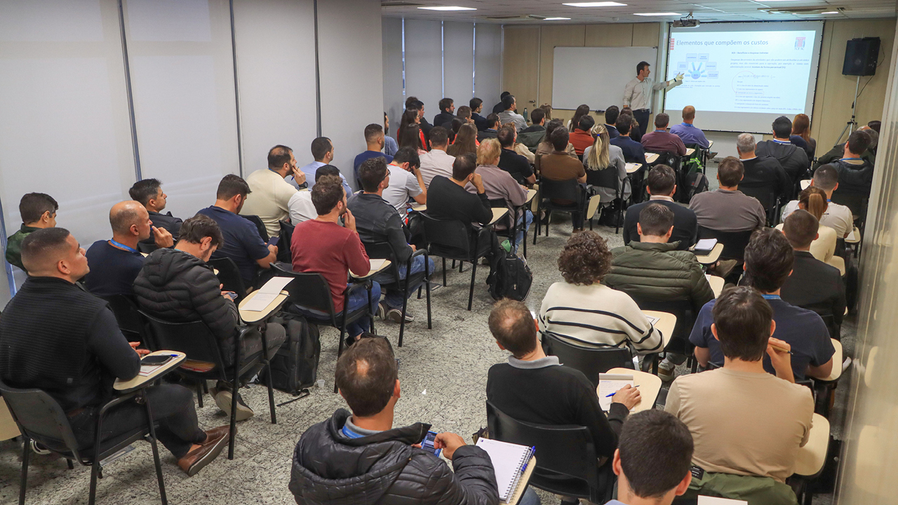 Imagem mostra várias pessoas de costas, sentadas em carteiras em uma sala do TCE/SC. À frente deles, um dos técnicos faz apresentação. No telão, ao fundo, é projetado slide.