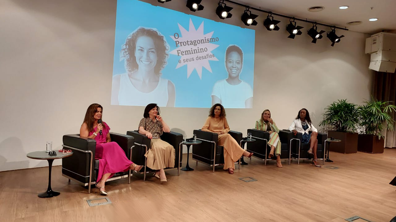 Foto de cinco mulheres sentadas em poltronas no palco do auditório do evento. Entre elas, a procuradora-geral-adjunta Cibelly Farias. Ela é uma mulher branca, de cabelos castanhos. Usa um vestido rosa. Ao fundo, projeção, de uma apresentação.