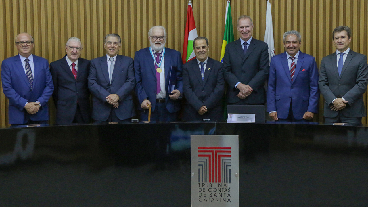 Foto do plenário do TCE/SC onde aparecem, atrás da bancada, em pé, da esquerda para a direita, os conselheiros Wilson Wan-Dall, Moacir Bértoli, César Fontes, Salomão Ribas Jr., Otávio Gilson dos Santos, Herneus De Nadal, Aderson Flores e José Nei Ascari.