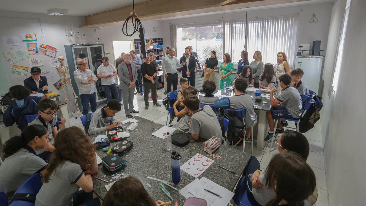 Um grupo de pessoas aparece em pé, ao fundo, em uma sala de aula. Eles estão de frente para um grupo de crianças.