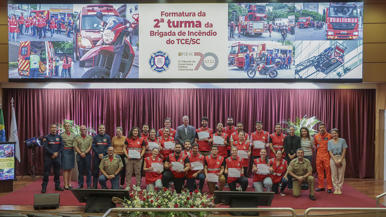 Banner horizontal com foto dos 15 novos formados, vestindo uniforme vermelho, lado a lado e junto com o presidente do TCE/SC, conselheiro Herneus De Nadal, e autoridades do Corpo de Bombeiros e da Polícia Militar. Eles estão no palco do auditório bordô do TCE/SC. No telão, na parte superior, banner com imagens de bombeiros.
