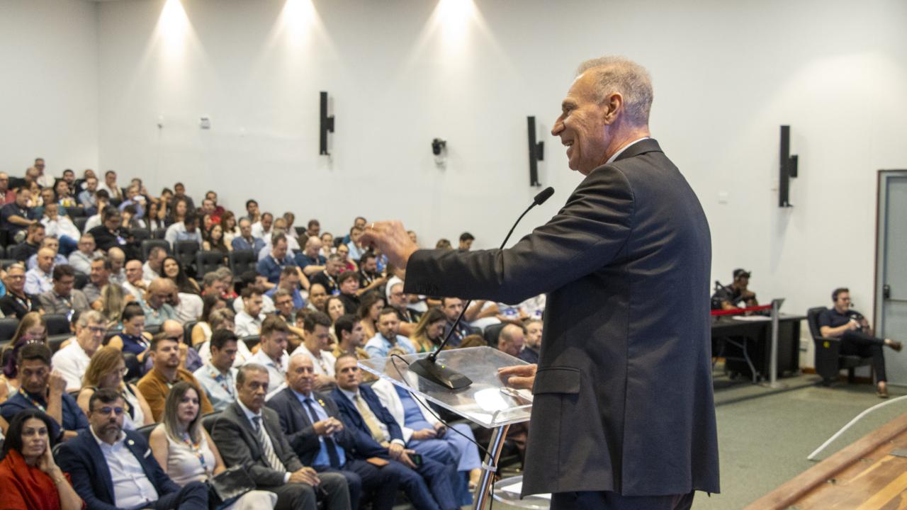 Imagem mostra em primeiro plano um homem falando ao microfone e vestindo um paletó escuro. Ao fundo, há uma plateia olhando para ele