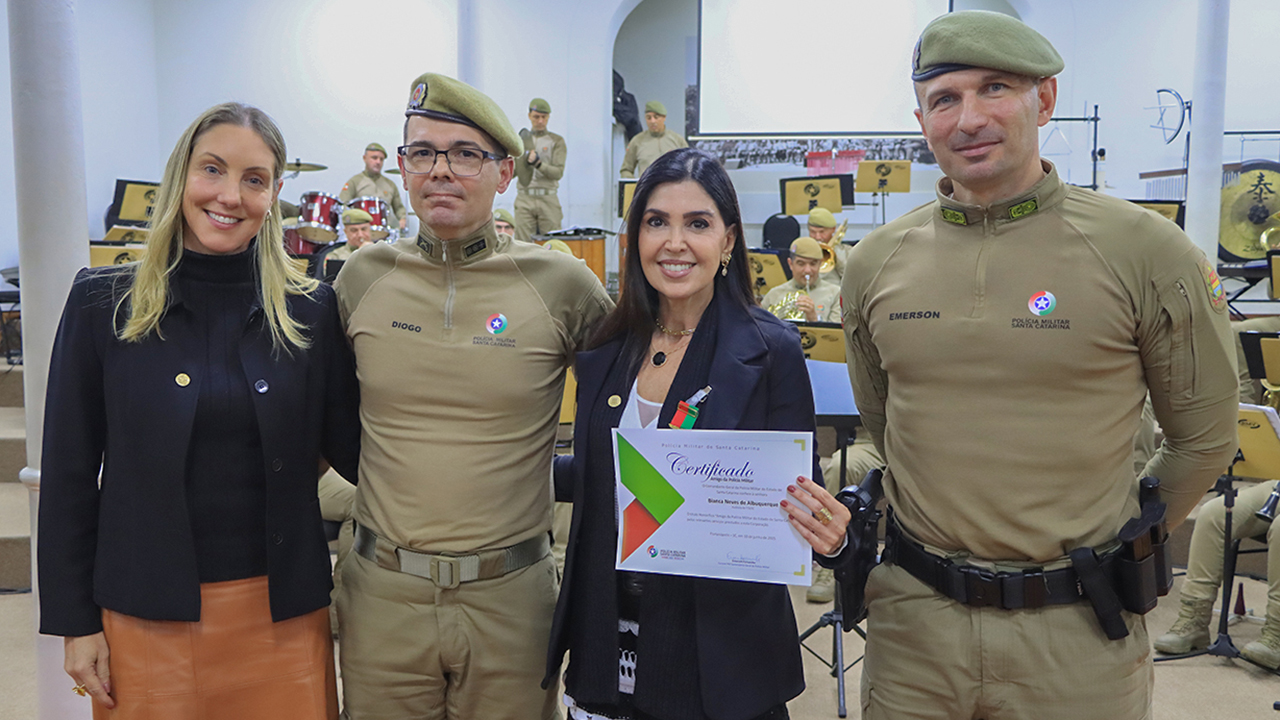 Banner horizontal com foto de quatro pessoas, lado a lado, durante a solenidade. Há dois servidores da PMSC, a auditora fiscal de Controle Externo Bianca Albuquerque e a diretora-geral de Administração do TCE/SC, Thais Serpa. Todos sorriem para a foto. Bianca segura o certificado com a homenagem recebida.