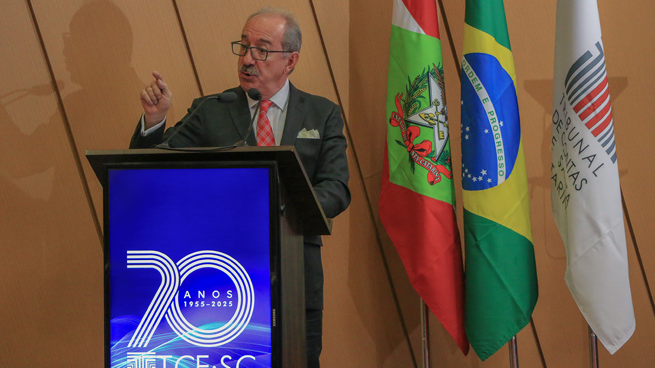 Foto horizontal do palestrante José Juis Garcia Guerrero que está falando do púlpito. Ele é um homem branco, usa óculos e veste um terno cinza escuro. Atrás dele, as bandeiras de Santa Catarina, do Brasil e do TCE/SC.