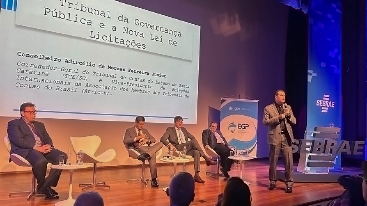Foto do auditório onde o conselheiro Adircélio, a frente, de pé e com microfone, faz palestra no evento. Ao fundo, sentados em quatro cadeiras, estão outros expositores e painelistas. Na parede do fundo, é projetada a apresentação realizada pelo conselheiro Adircélio. No canto direito do palco, um banner com a logomarca do evento.