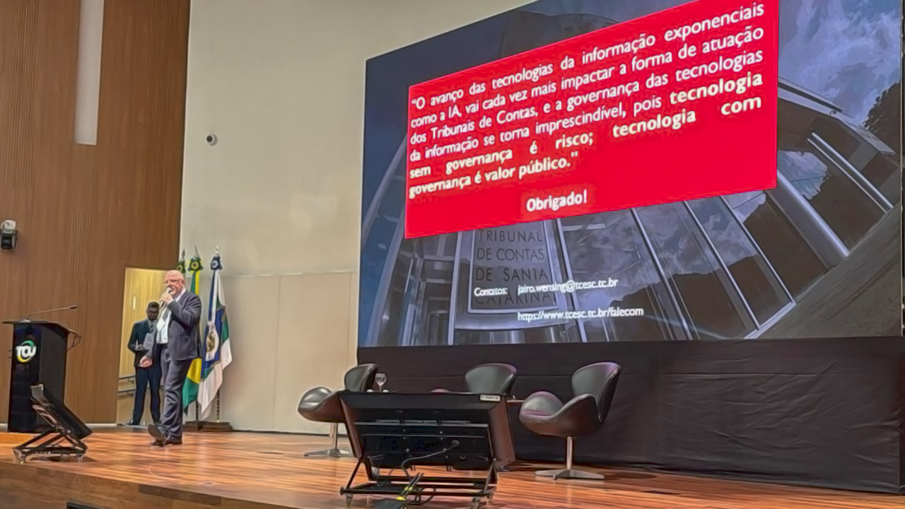 Banner horizontal com foto do assessor da Aget, Jairo Wensing, falando ao público durante apresentação no Enastic Controle 2025, no auditório do Instituto Serzedello Corrêa. Ele está em pé, no palco. Ao fundo, no telão, é exibida a sua apresentação. 