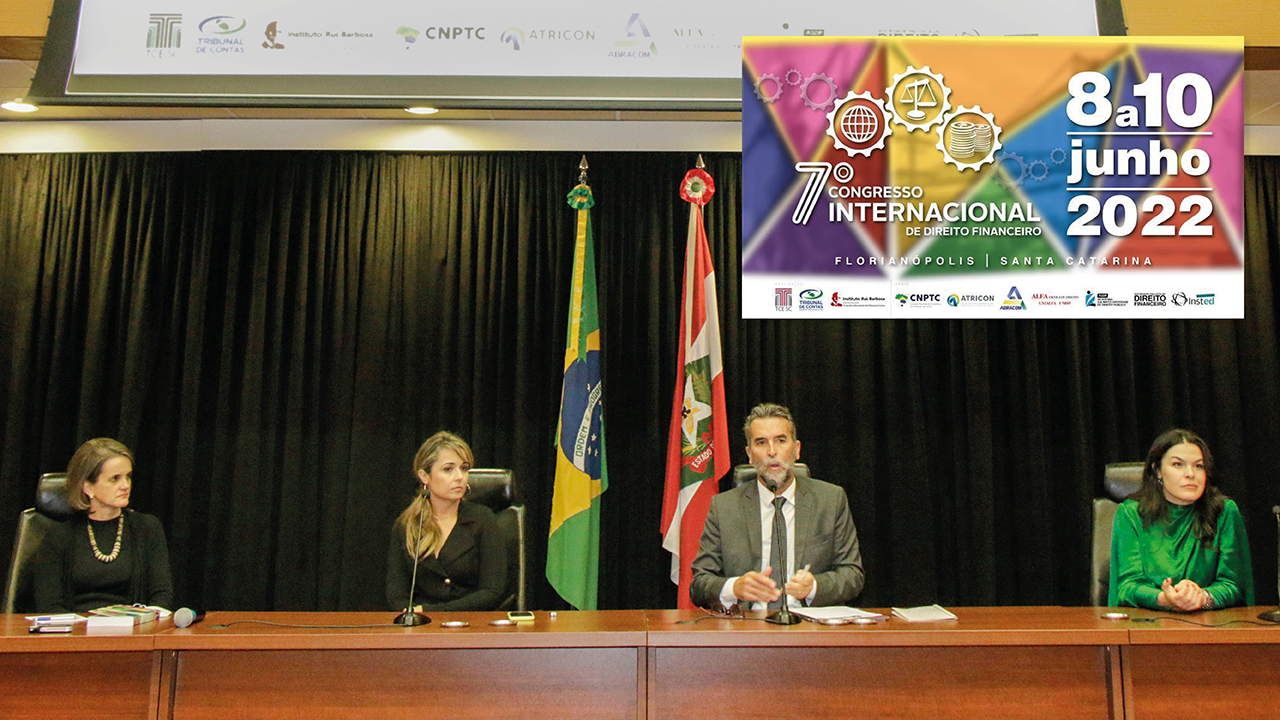 Da esquerda para direita as palestrantes Paula Schommer e Gissele Nunes, o mediador Marcelo Brognoli da Costa e a palestrante Monique Porte sentados à mesa. Ao fundo uma bandeira do Brasil e outra de Santa Catarina