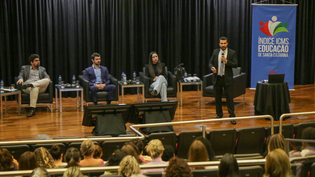   Foto do palco do TCE/SC. Nele estão quatro pessoas: 2 homens sentados, 1 em pé. Uma mulher sentada à direita. Em primeiro plano, na parte inferior da imagem, pessoas, de costas, na plateia.