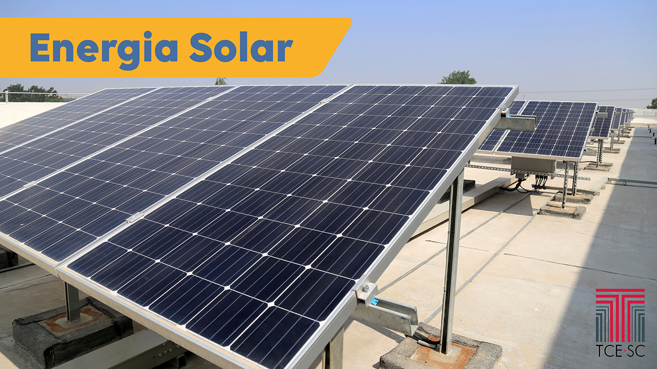 Imagem mostra em primeiro plano placas de coleta de energia solar, os painéis fotovoltaicos. No alto, à esquerda, sobre fundo amarelo, está escrito em azul a expressão energia solar.