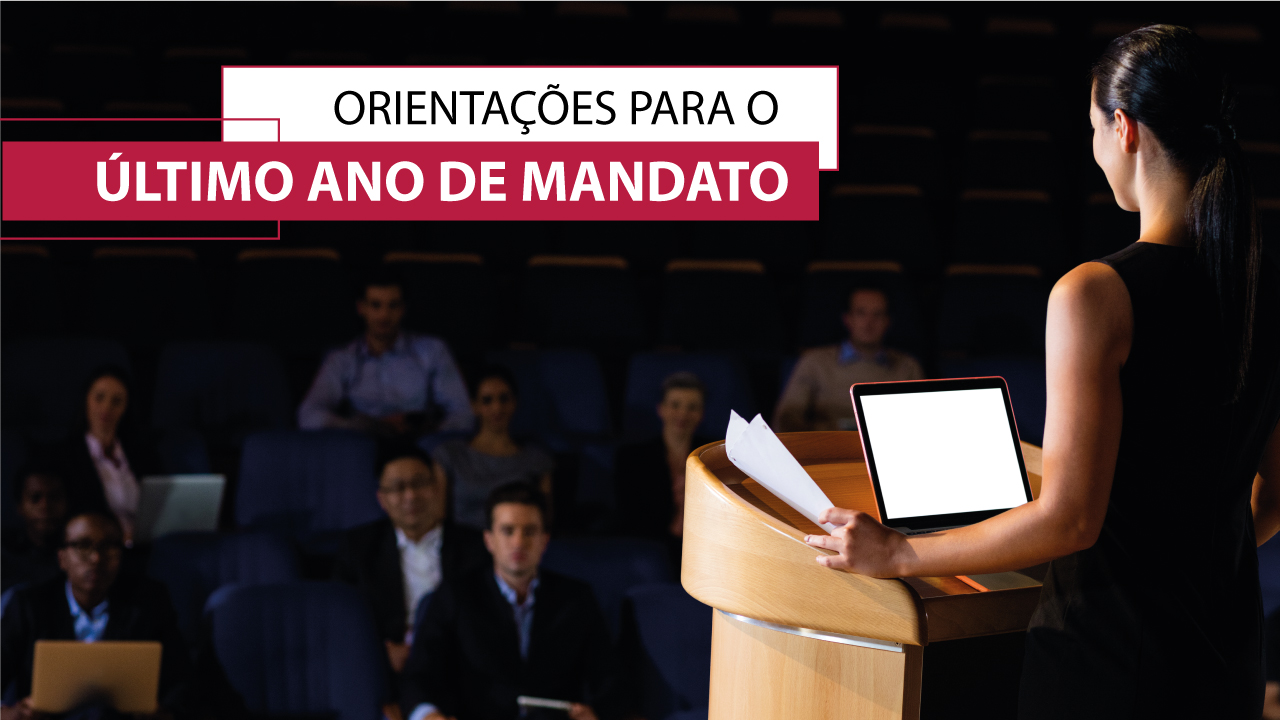 Imagem mostra em primeiro plano, à direita, uma mulher em pé falando em um púlpito, onde há um computador ligado. Ao fundo, aparece a plateia em um auditório. No alto, mais centralizado, há a informação “Orientações para o último ano de mandato”