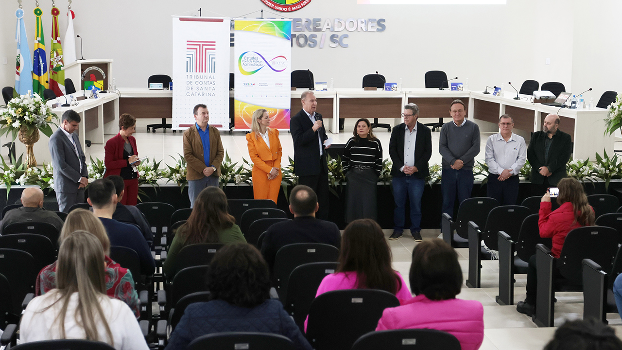 Foto do presidente Herneus De Nadal falando ao público durante a segunda etapa do 23º Ciclo de Estudos, em Palmitos. Ao lado dele, em pé, estão outras nove pessoas. Ao fundo, há o banner com o logo do TCE/SC e do Ciclo de Estudos. À sua frente, há o público que o assiste, de costas para a foto, sentado em poltronas.