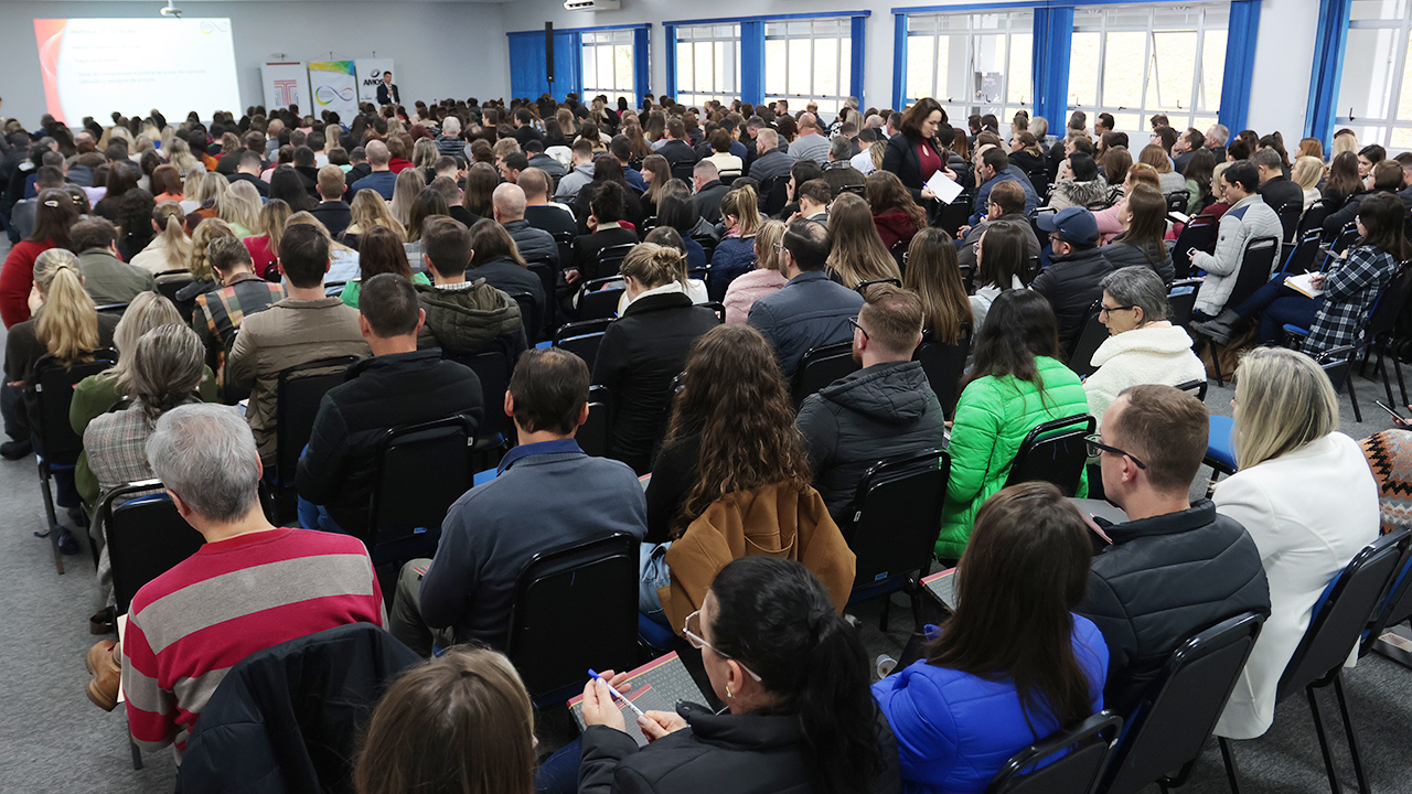 Foto do auditório lotado durante a etapa de Chapecó do Ciclo de Estudos. Há várias pessoas, entre homens e mulheres, de costas para a foto, sentadas em cadeiras pretas, assistindo à uma das falas. 