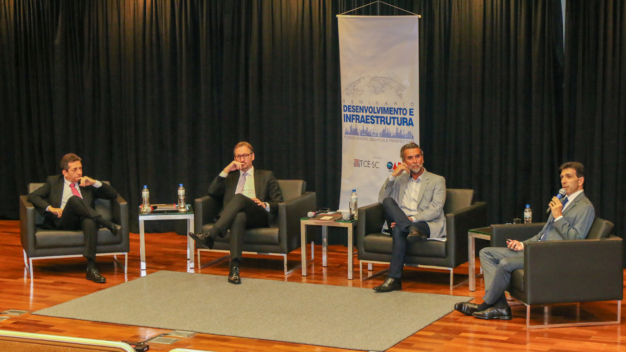 Imagem mostra os palestrantes em um sentados em cadeiras no palco. O representante do TCE/SC, Rogério Loch, está à direita da imagem, com um microfone na mão