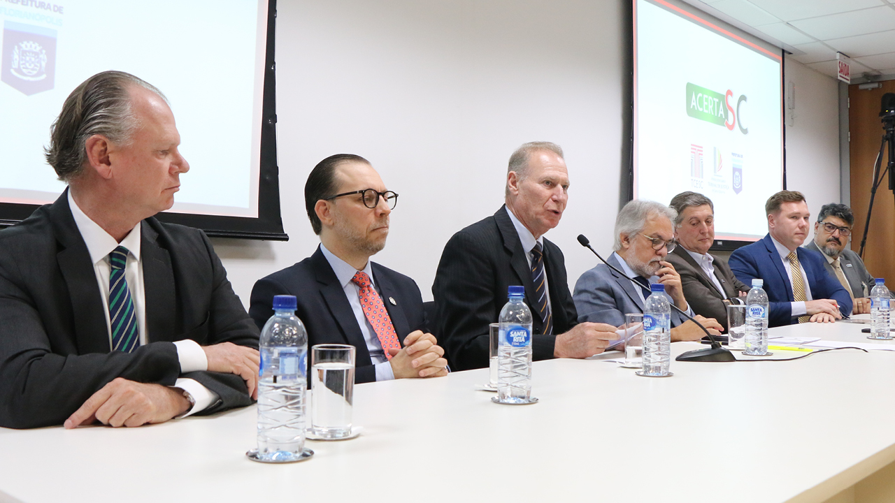 Imagem mostra sete homens sentados à mesa. O conselheiro Herneus De Nadal, o terceiro da esquerda para a direita, está falando ao microfone. Sobre há mesa há garrafas plásticas de água e copos.