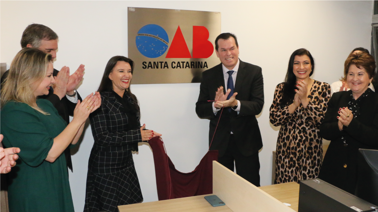 Foto mostra uma placa da OAB/SC com o presidente do TCE/SC, Adircélio de Moraes, e a presidente da OAB/SC, Cláudia Prudêncio, sorrindo em frente à placa.