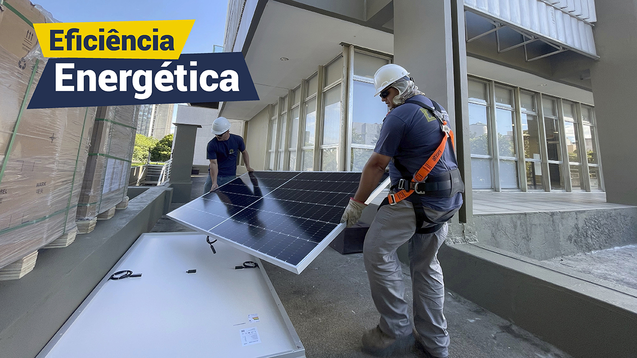 Imagem ao fundo prédio do Tribunal de Contas. No primeiro plano dois homens brancos vestindo uniformes azuis colocam no chão painéis solares.