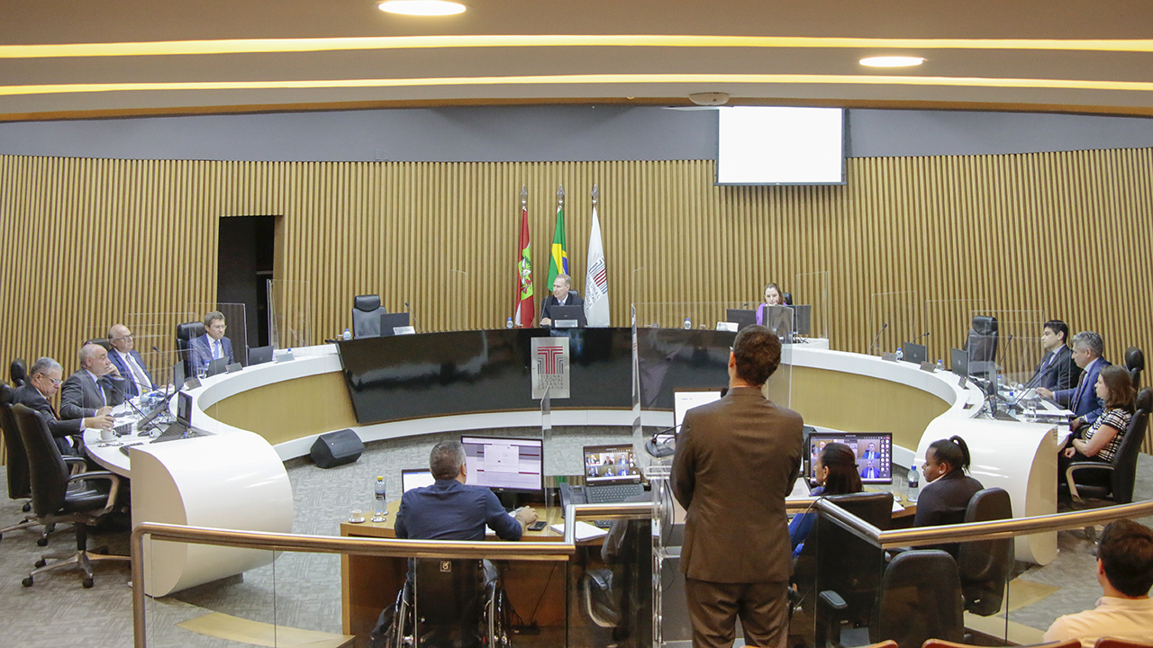 Imagem mostra o plenário do TCE/SC. O fundo é em tom amarelado. Dispostos em uma mesa que acompanha a parede arredondada estão os conselheiros. Em primeiro plano, de costas, aparecem as pessoas que compõem o secretariado do Pleno.