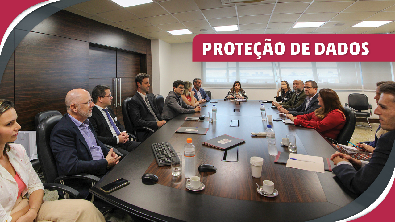 Imagem mostra a fotografia de um grupo de pessoas em reunião ao redor de uma mesa. No canto superior direito, em destaque e em letras brancas sobre fundo vermelho, lê-se "Proteção de Dados".