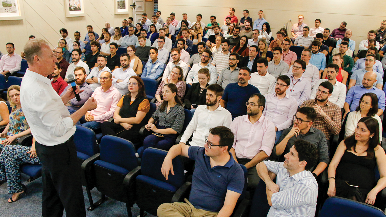 Presidente do TCE/SC, Herneus De Nadal, está em pé, à esquerda, falando ao microfone. Demais pessoas estão sentadas à sua frente.  Há cerca de 60 pessoas na imagem.