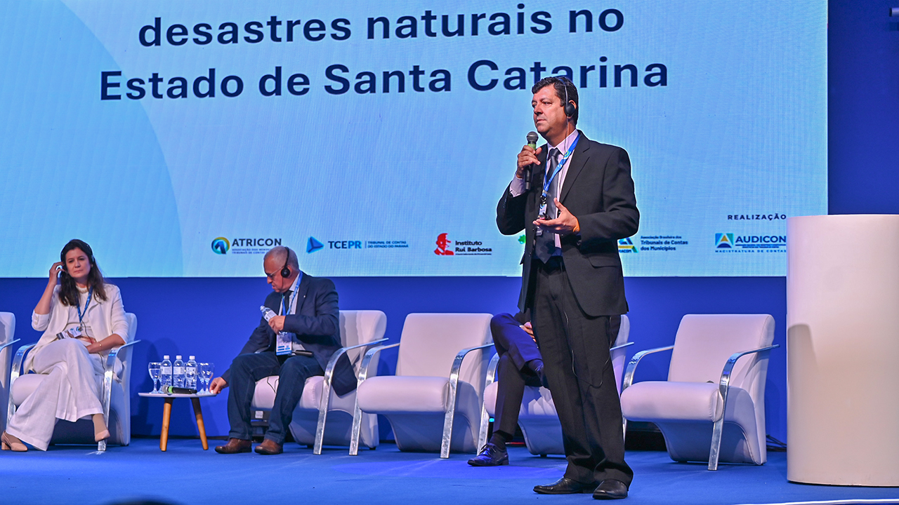 Foto do auditor da DAE do TCE/SC Osvaldo Faria de Oliveira. Ele, um homem branco de terno escuro, está ao lado esquerdo da foto. Atrás dele, no palco, um homem e uma muher estão sentado. Ao fundo um telão com uma imagem onde está escrito "desastres naturais no Estado de Santa Catarina.