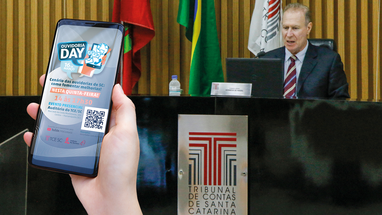 Foto do plenário do TCE/SC, com o presidente Herneus De Nadal, de terno escuro e gravata listrada, em sua posição na mesa, vendo-se ao fundo as bandeiras de Santa Catarina, do Brasil e do TCE/SC e a logo do TCE/SC na parte frontal da tribuna. Em primeiro plano, à esquerda, uma mão segura um celular com a imagem de divulgação do evento “Ouvidoria Day” na tela.