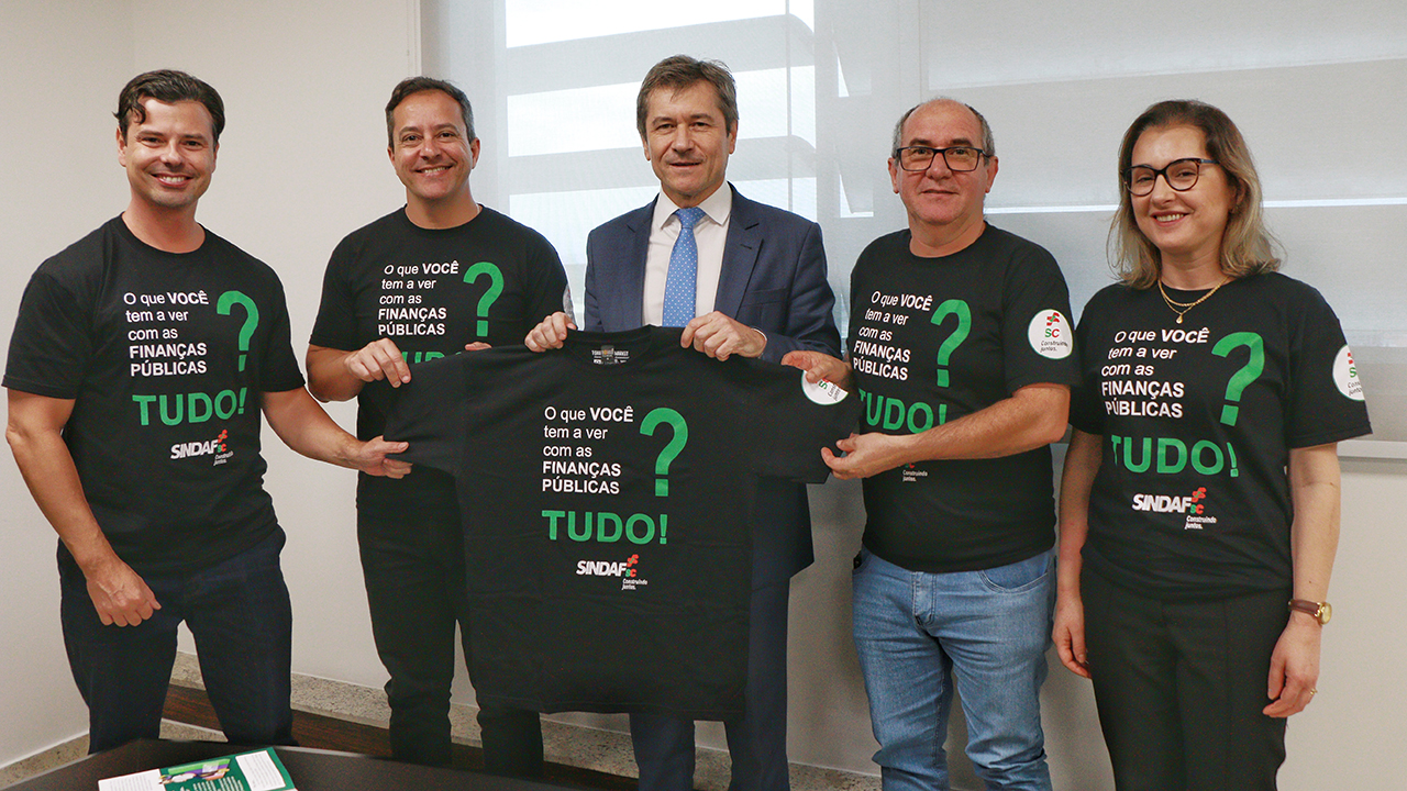 Foto dos participantes da reunião entre o TCE/SC e o Sindaf/SC. Há cinco pessoas em pé, uma ao lado da outra, segurando uma camiseta preta que contém a frase “O que você tem a ver com as finanças públicas? Tudo!”. Entre elas, o conselheiro José Nei Ascari, que veste terno e gravata, e os membros do Sindaf, que vestem camisetas iguais a que seguram. 