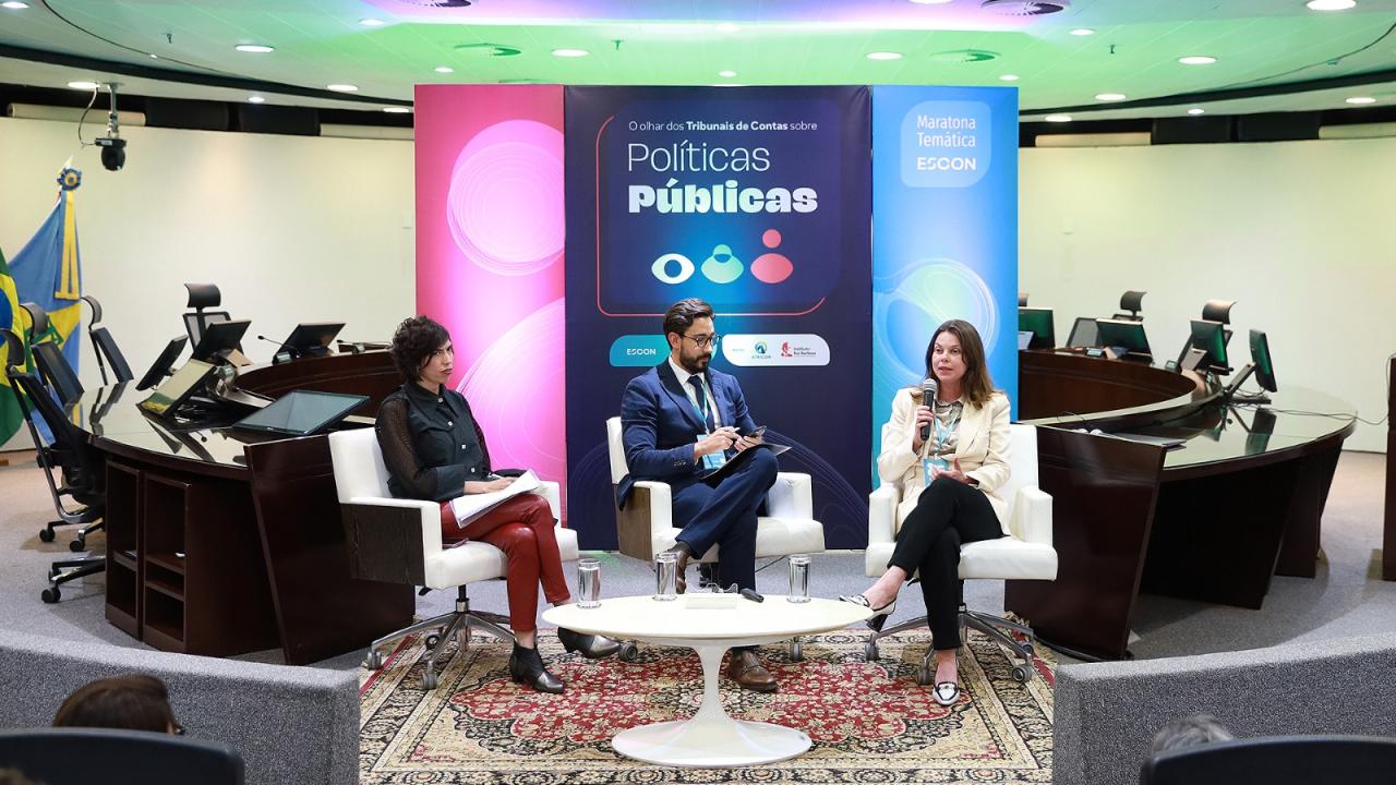 Foto de três painelistas durante o evento. Entre os presentes, a conselheira substituta do TCE/SC Sabrina Iocken e a professora da FGV Gabriela Lotta. Ao fundo, um telão exibe a apresentação intitulada “Políticas públicas”. Sabrina fala ao microfone. Os três estão sentados em poltronas no palco. 