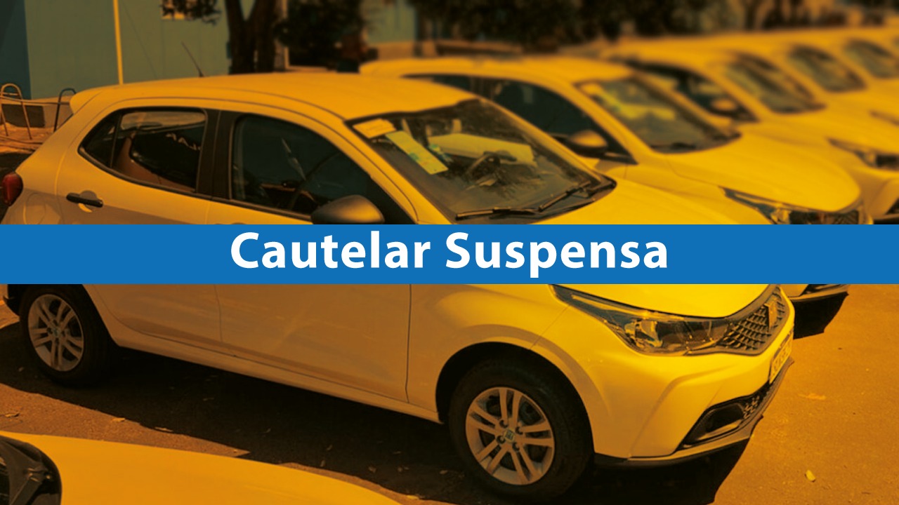 Imagem de uma fileira de carros compactos estacionados, todos na cor branca, vistos em um pátio ao ar livre. Sobre o carro em primeiro plano há uma faixa horizontal azul com o texto em branco “Cautelar Suspensa” 