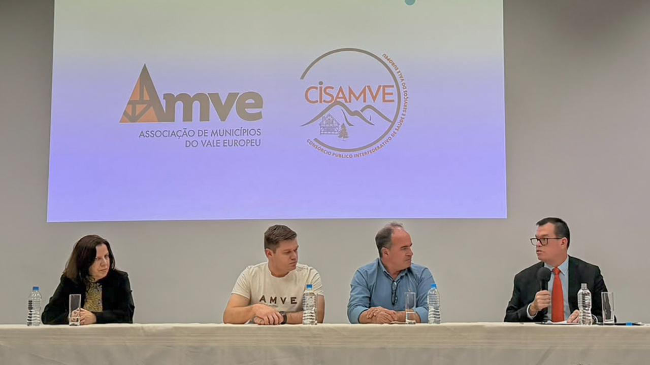 Foto horizontal da mesa composta do evento. À esquerda, o conselheiro e Corregedor-Geral do TCE/SC Adircélio de Moraes Ferreira Júnior. Ao lado outras 3 pessoas - 2 homens e uma mulher. Ao fundo projetada a logomarca da AMVE.