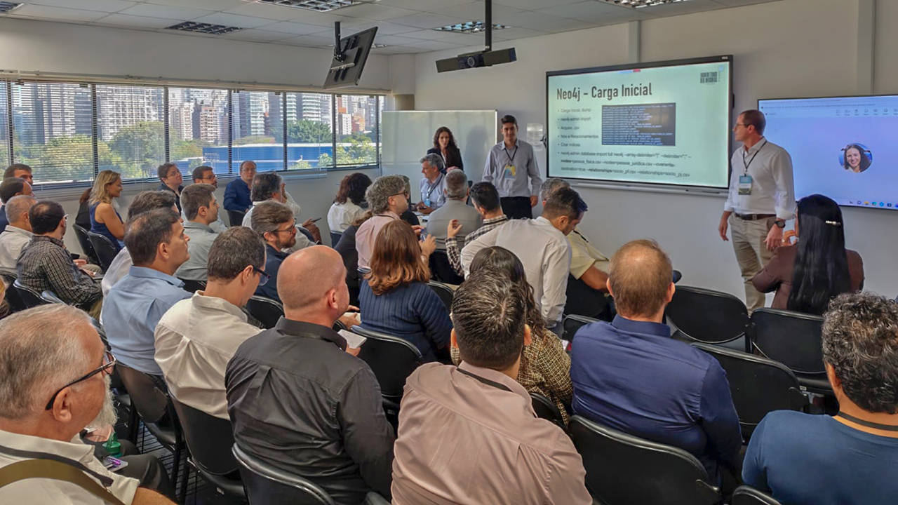 Banner horizontal com foto de uma sala de treinamento lotada durante o Infocontas 2026, com dezenas de participantes sentados em cadeiras e atentos a uma apresentação técnica. À frente do ambiente, os auditores fiscais de Controle Externo Nilsom Zanatto, Roberto Fleischmann e Thiago Sodré apresentam o Sistema Visão, posicionados próximos a uma tela de projeção que exibe slides sobre o tema. Os participantes observam a exposição, alguns fazendo anotações, enquanto os palestrantes interagem com o público. 
